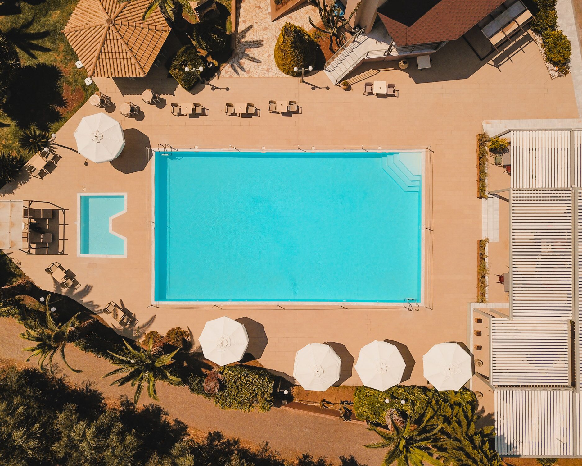 Vista aérea de un hotel con una gran piscina rectangular, zonas de estar al aire libre y palmeras.
