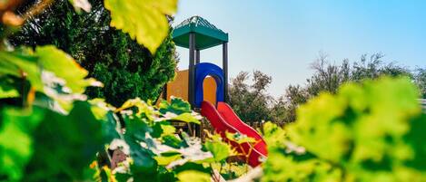 Un terrain de jeux avec un toboggan et une structure d’escalade, le tout entouré de verdure.