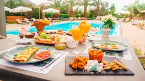 A table set with breakfast items including pancakes, eggs, and fruit, with a pool view in the background. - Apollo Resort Art Hotel (Trifylia)