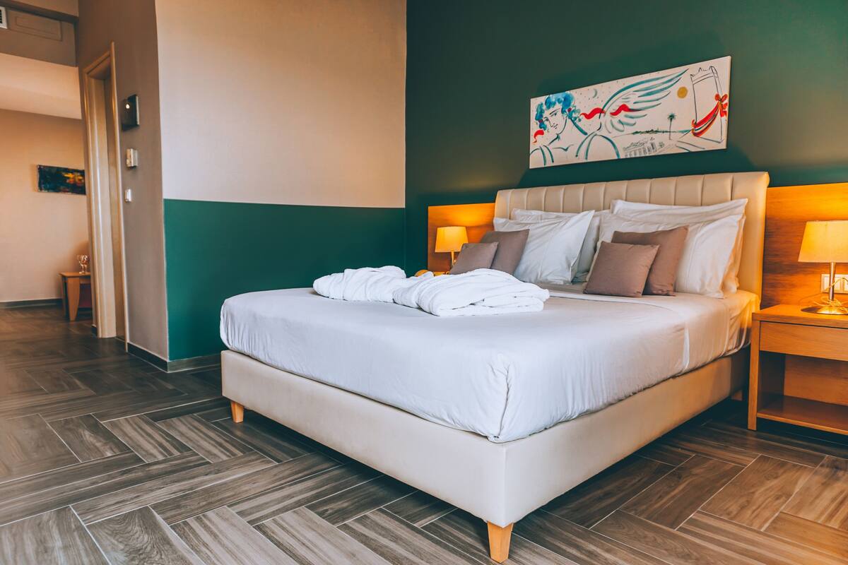 A neatly made bed with white linens and pillows, a headboard with a decorative painting, and a wooden floor.