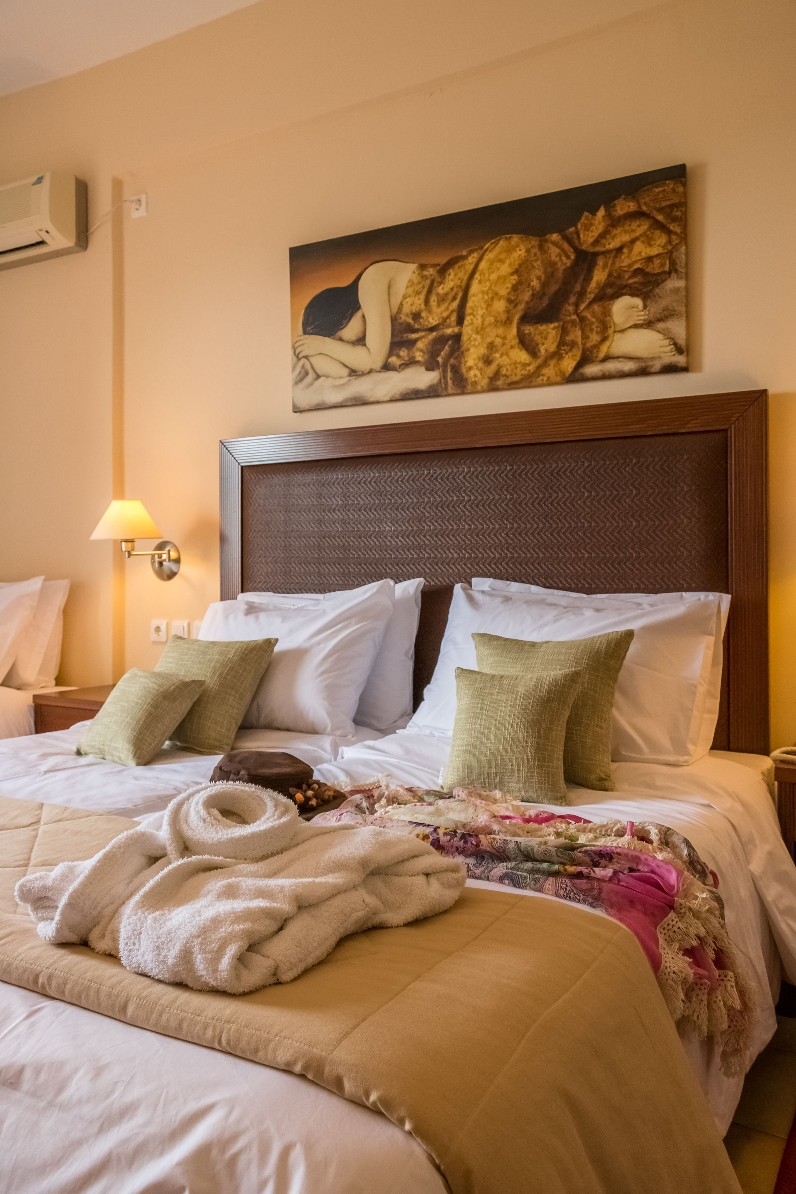 A hotel room with a large bed, a headboard, a painting above the bed, a nightstand with a lamp, and a wall-mounted air conditioner.
