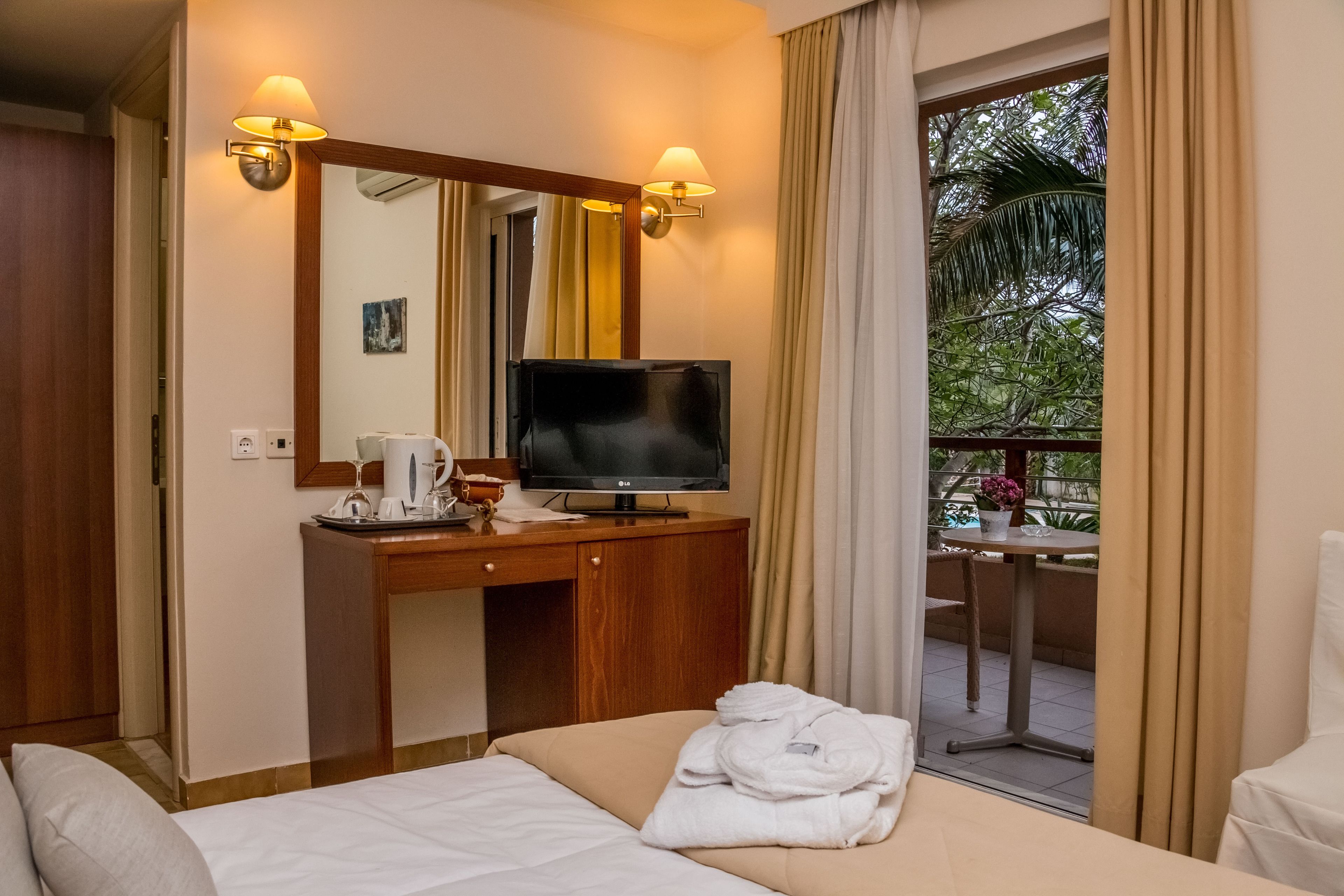 A hotel room with a bed, a TV on a wooden desk, a mirror, and a view of the outdoors.