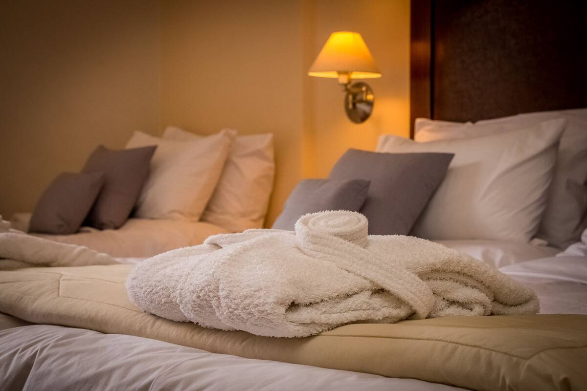 A neatly made bed with white and gray pillows, a rolled white bathrobe, and a wall-mounted lamp.