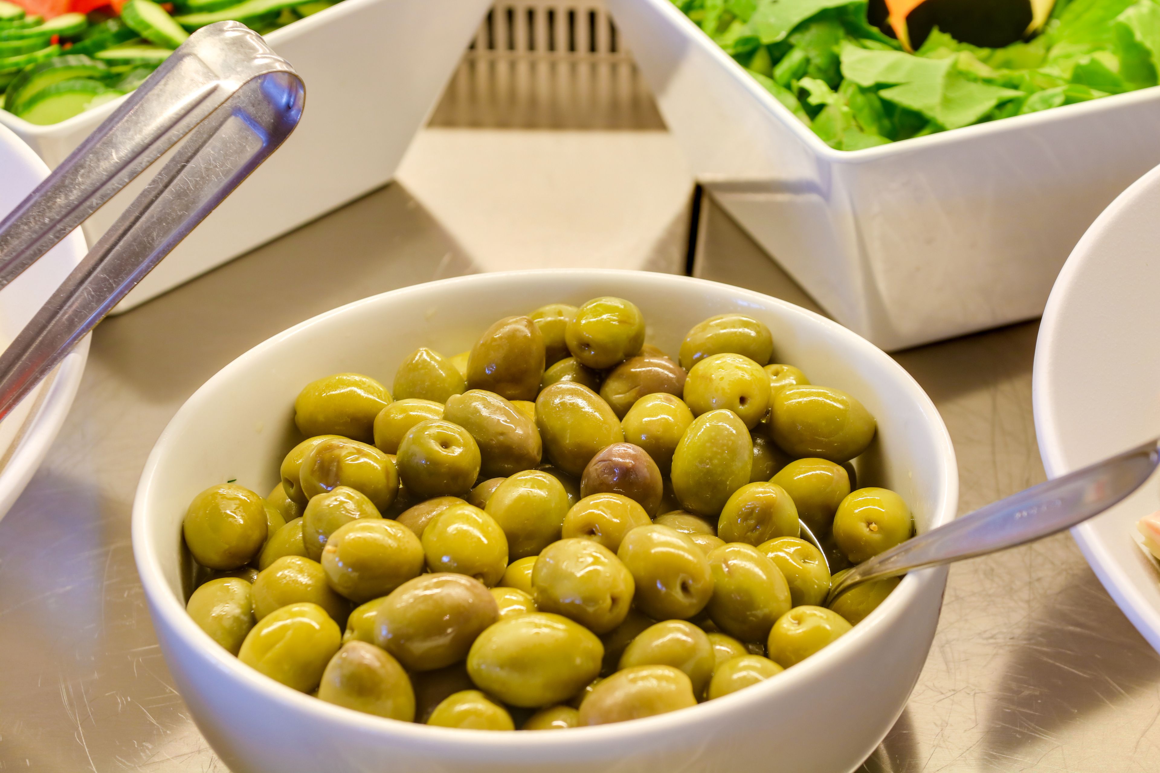A bowl of olives with a spoon, surrounded by salad containers.