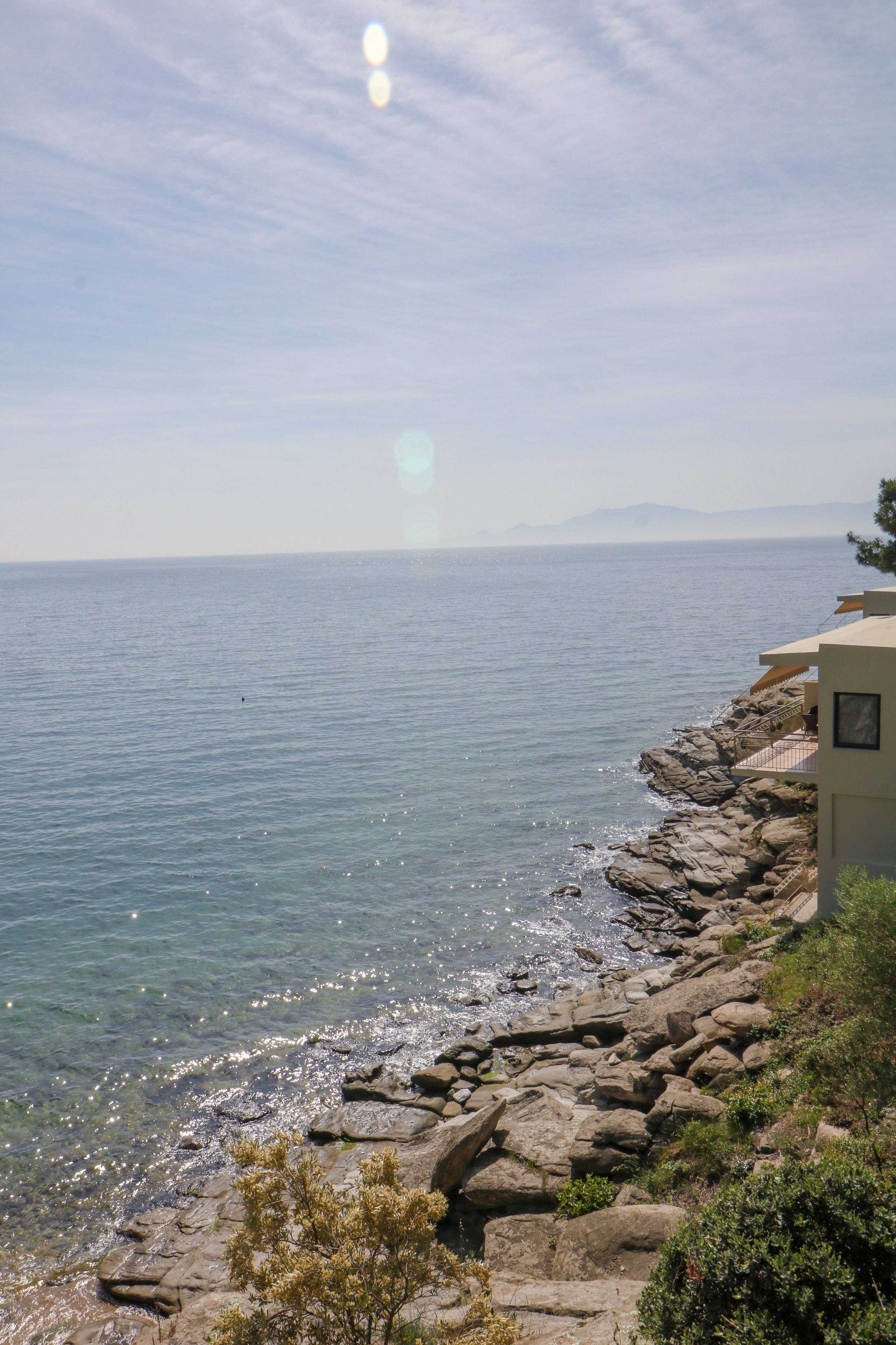 A coastal view with a rocky shoreline, clear sky, and a distant mountain range.