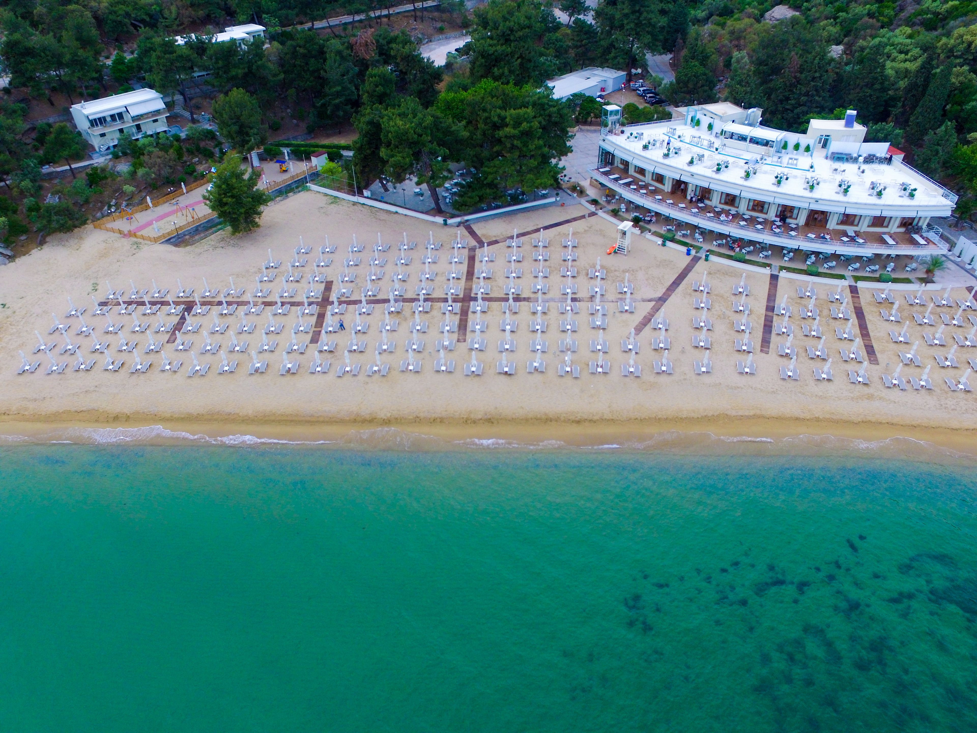 An aerial view of a beachside resort with a large number of sun loungers arranged in rows, a building with a curved roof, and a clear view of the turquoise sea.