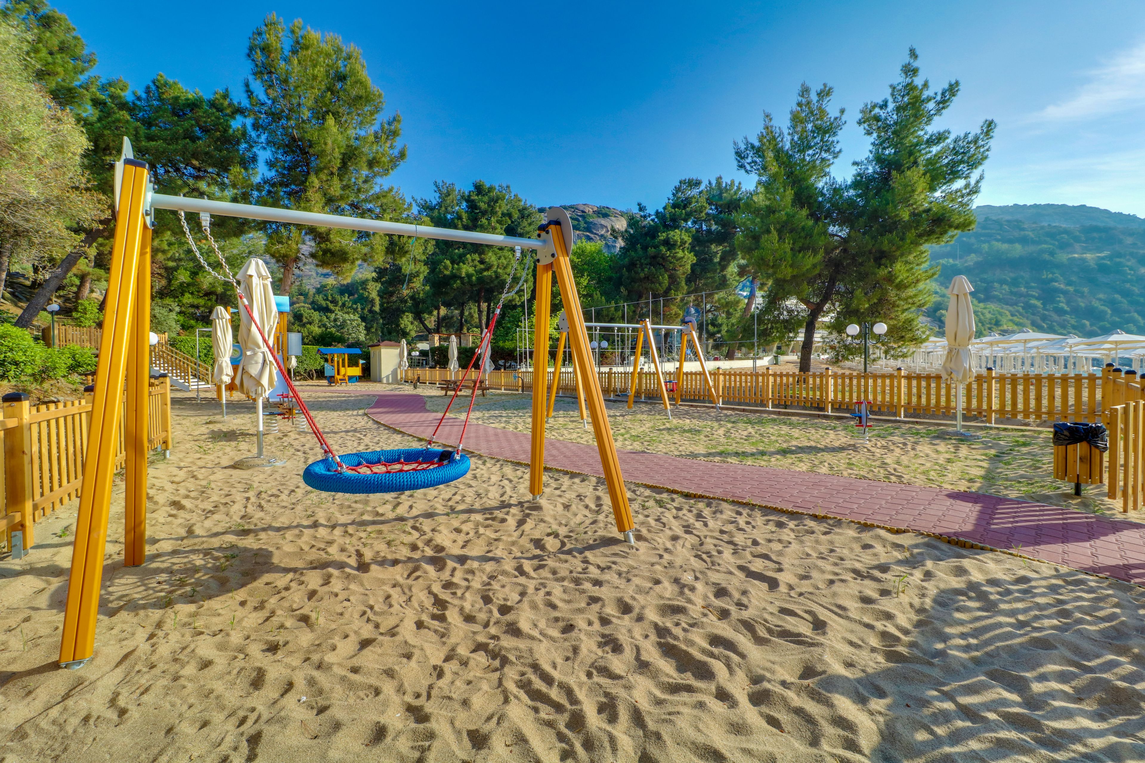 Ein Spielplatz mit Schaukeln und einer Tausschaukel auf Sandboden, umgeben von Bäumen und einem Holzzaun.