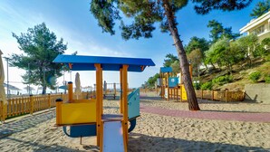 Zona arenosa de playa con juegos infantiles, incluyendo columpios y un tobogán, rodeada de árboles y una valla de madera.
