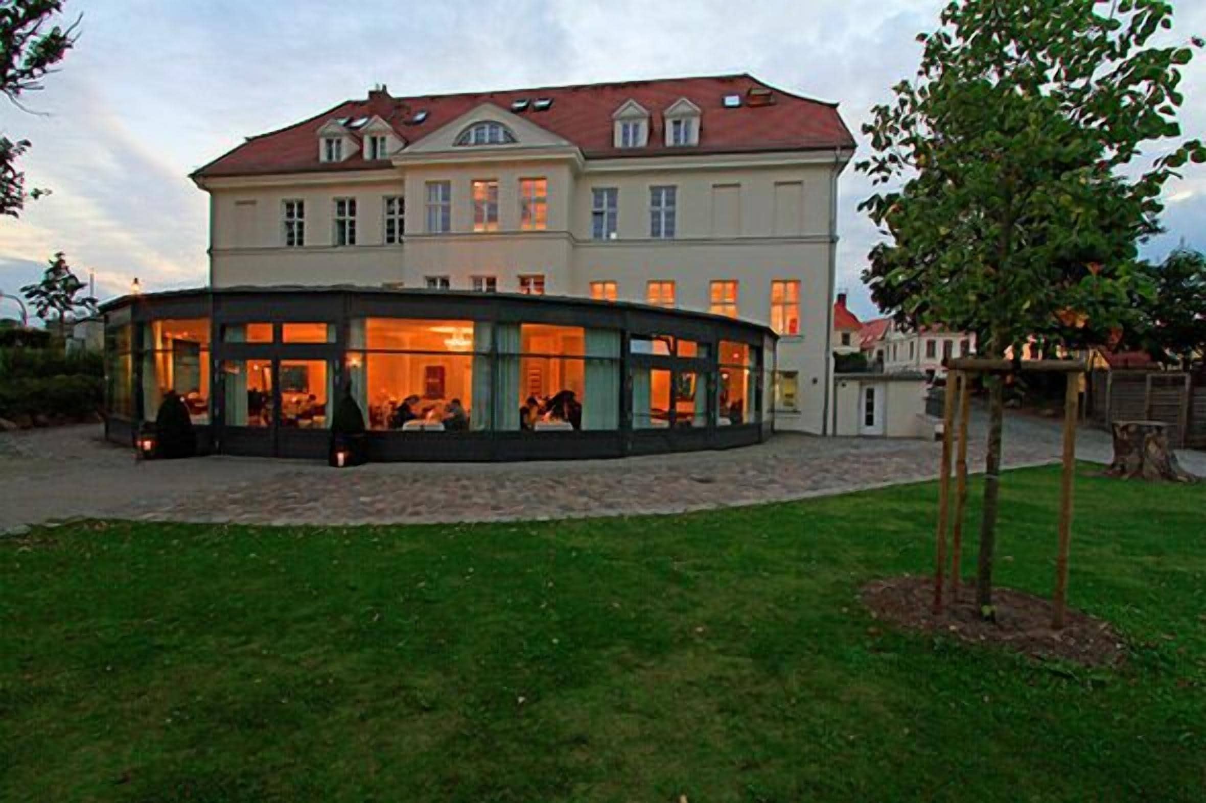 Un edificio grande con una estructura de vidrio curva al frente, probablemente un restaurante o cafetería, con un césped bien cuidado y un árbol cerca.
