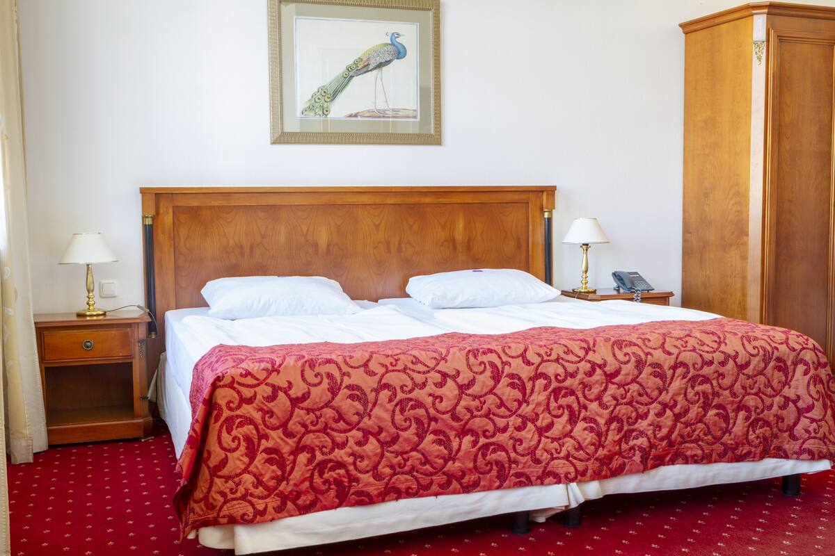 A hotel room with a wooden bed, two bedside tables with lamps, a telephone, and a peacock painting on the wall.