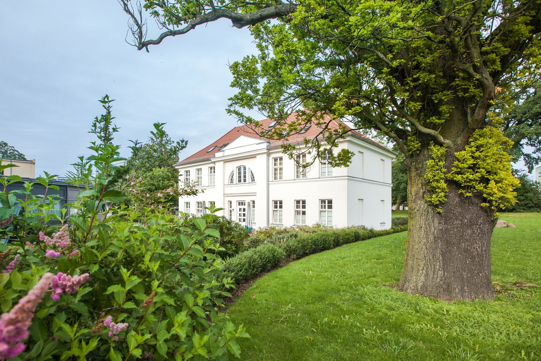 A white building with a red roof, surrounded by lush greenery and a large tree.