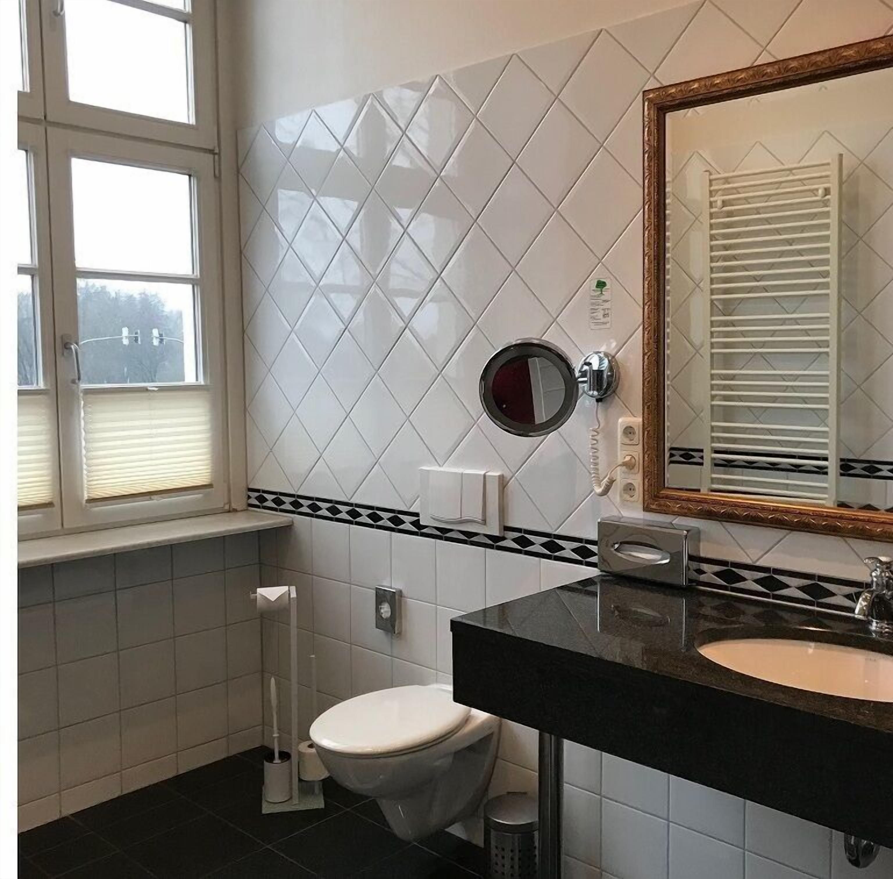 Baño con inodoro, lavamanos y espejo. Hay una ventana con persianas, dispensador de toallas y soporte para papel higiénico.
