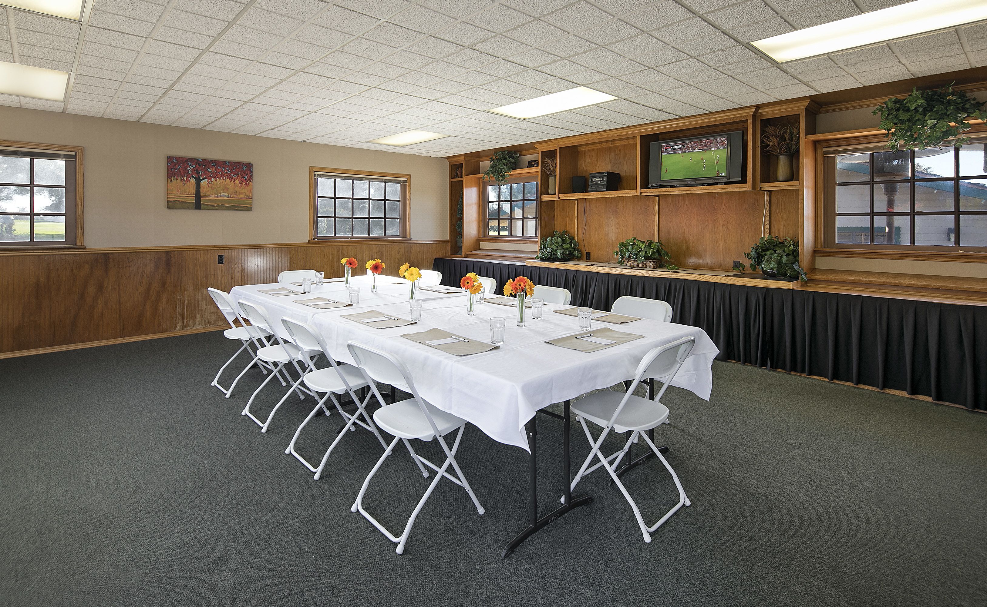 A conference room with a long table set for meetings, white folding chairs, and a television on a wooden shelf.
