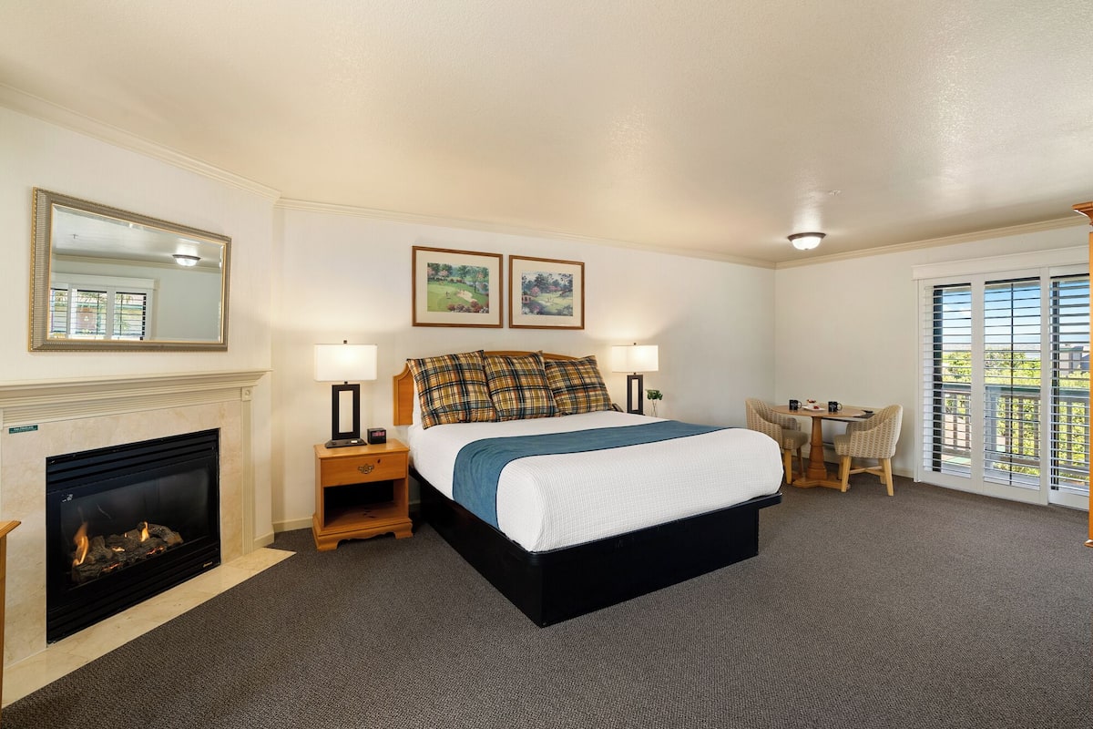 A hotel room with a bed, bedside lamps, a fireplace, a dining table with chairs, and two framed pictures on the wall.
