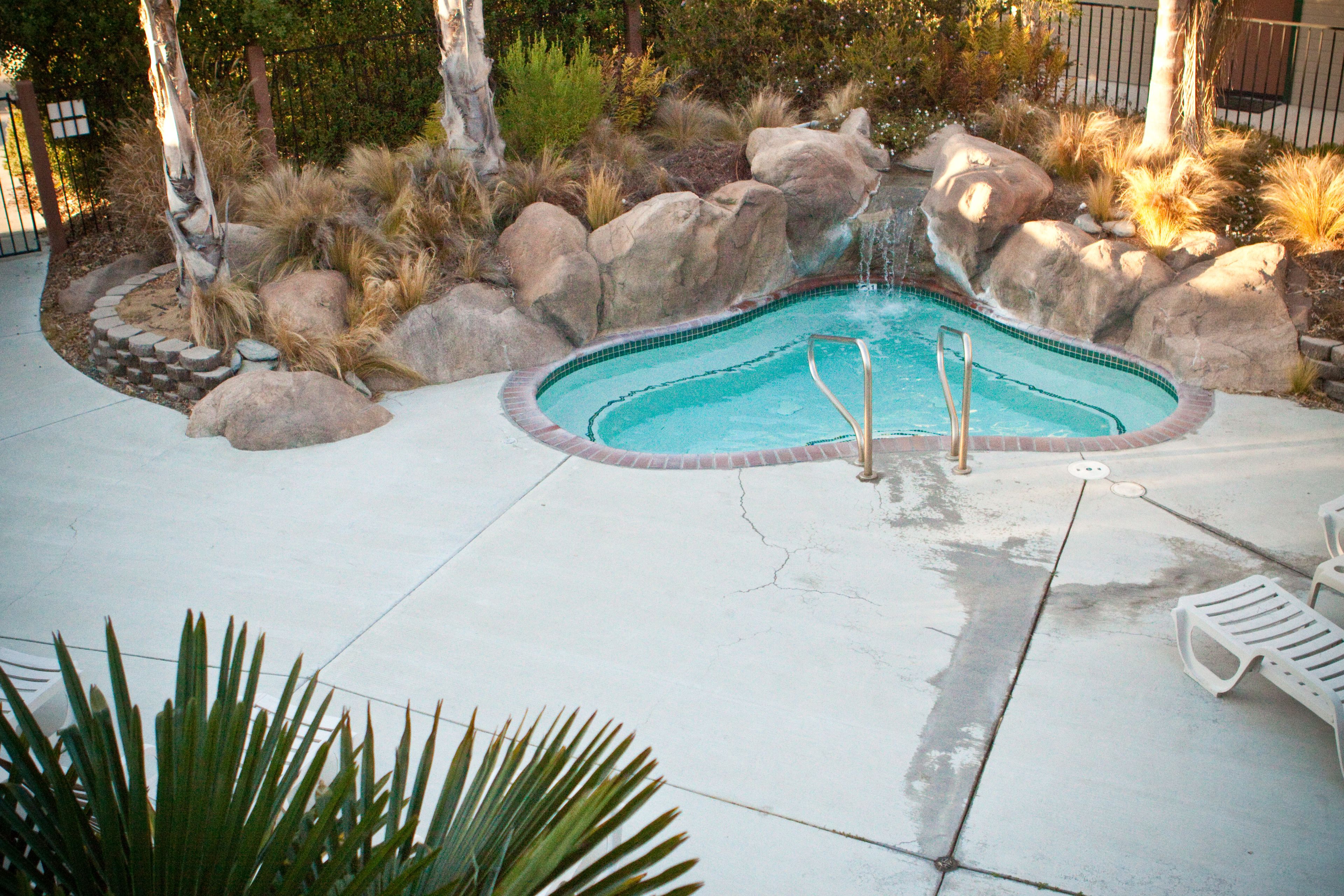 Una piscina circondata da rocce e piante, con un bordo in cemento e alcune sedie bianche di plastica.