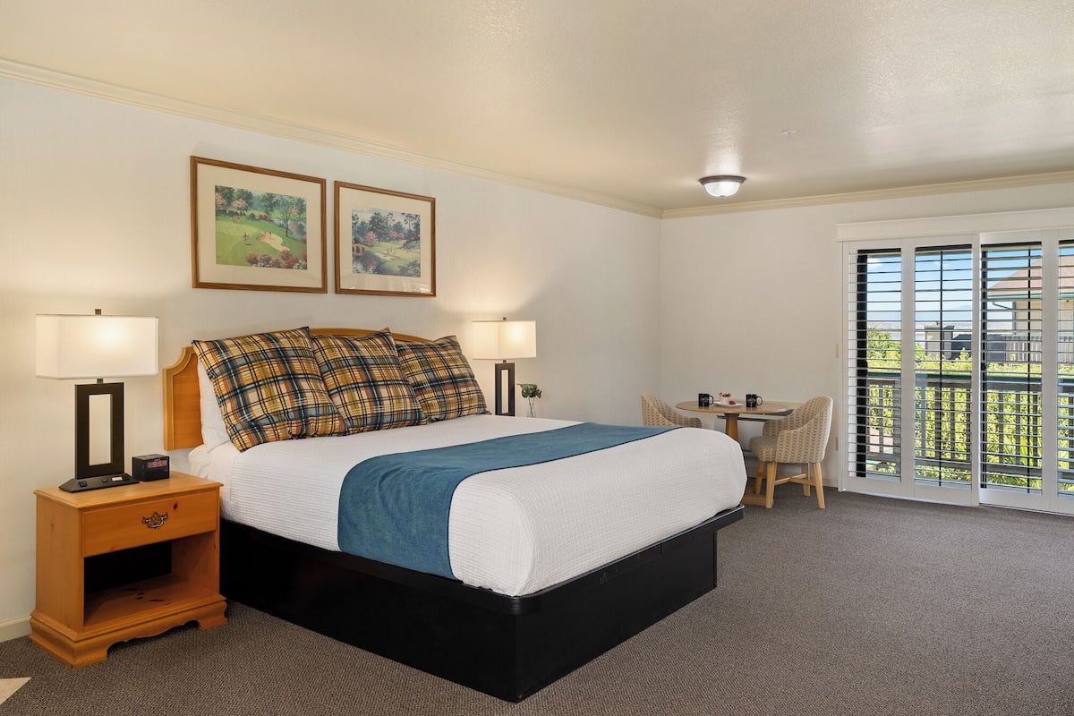 A hotel room with a bed, two bedside lamps, a nightstand, a small table with chairs, and a balcony with a view.