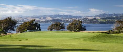 Un parcours de golf avec un drapeau, des arbres et une vue sur des montagnes et une étendue d’eau.