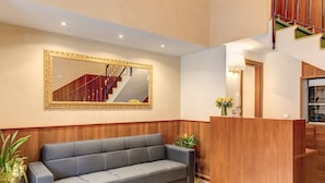 A reception area with a grey sofa, a wooden reception desk, a large mirror, and a staircase with wooden railings. - Hotel Flavio Rome (Rome)