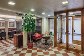 A lobby with a checkered floor, a large potted plant, a red sofa, a wooden reception desk, and a glass door entrance.