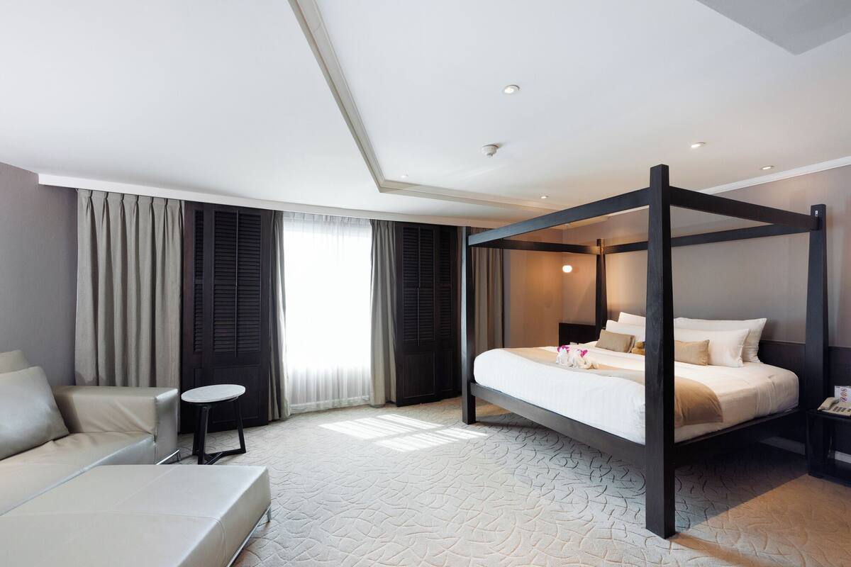 Une chambre d’hôtel moderne avec un lit à baldaquin, un canapé, une petite table ronde et une grande fenêtre avec des rideaux.
