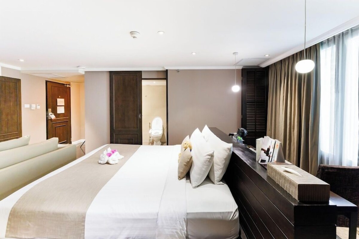 Habitación de hotel con una cama grande, una mesita de noche, un sofá y vistas al exterior por la ventana.