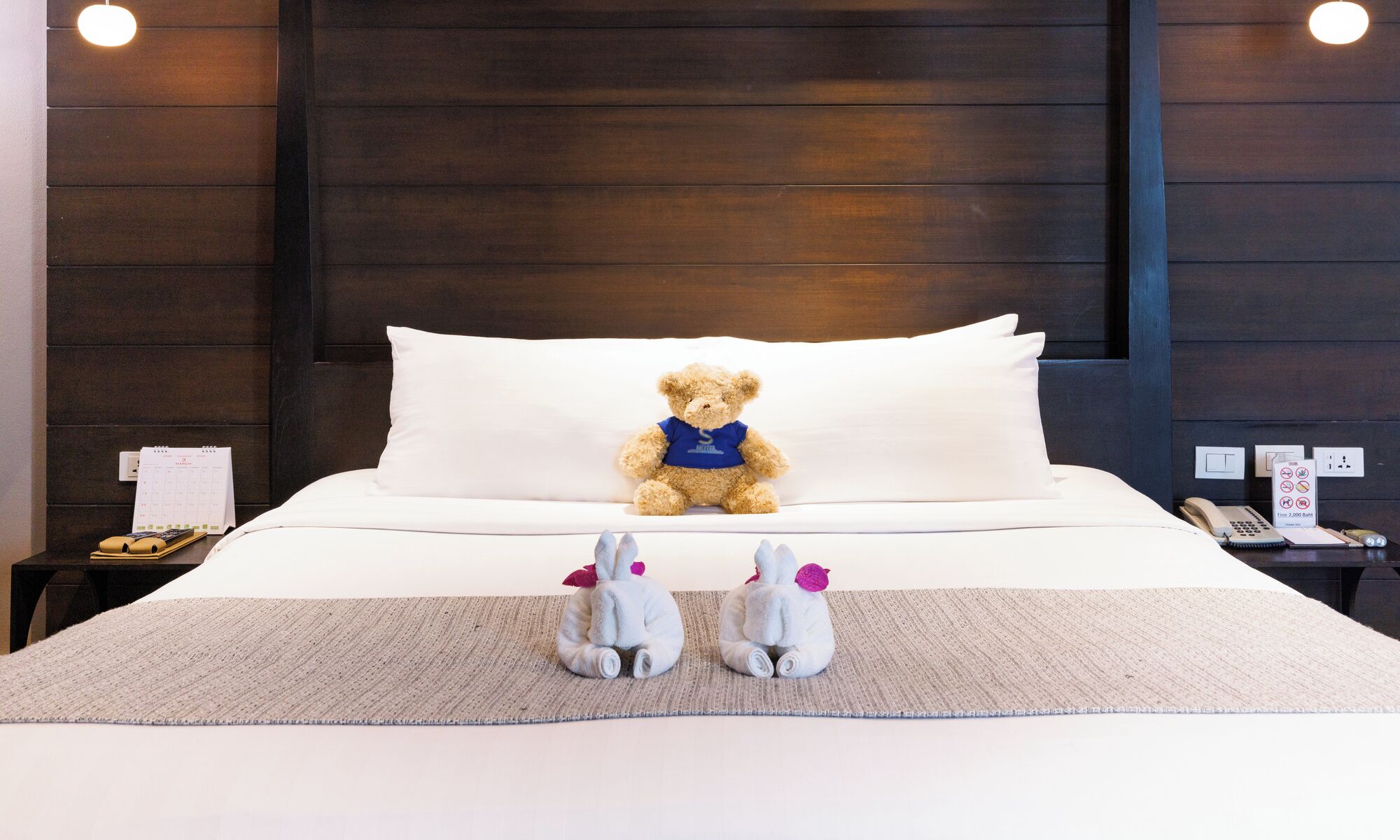A hotel room with a bed, a teddy bear on the pillow, a calendar, a phone, and a small table with a lamp.