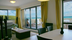 A modern room with a large sliding glass door leading to a balcony with a view of the ocean. There is a sofa, a coffee table, and a dining table with chairs. - Byala Beach Resort (Byala)