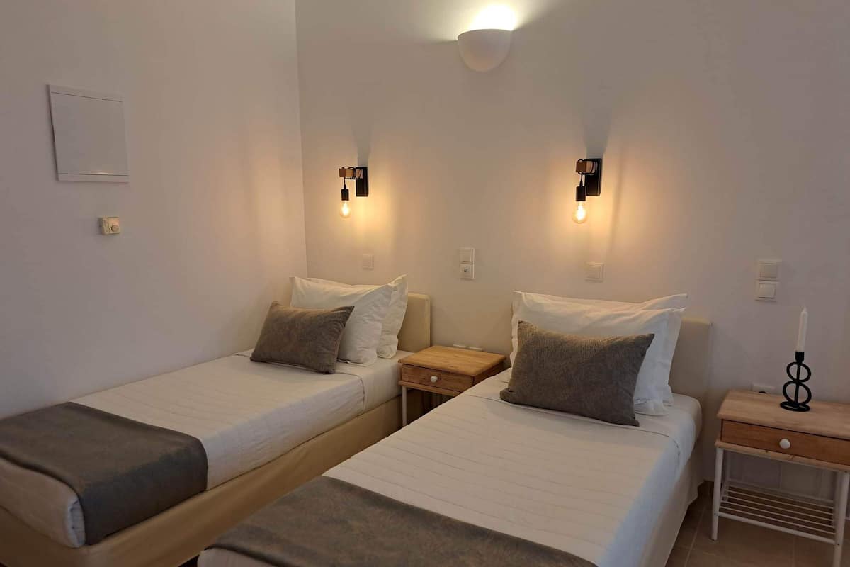 Habitación doble con dos camas individuales, cada una con una almohada y una manta. Hay mesitas de noche con lámparas y una lámpara de pared.
