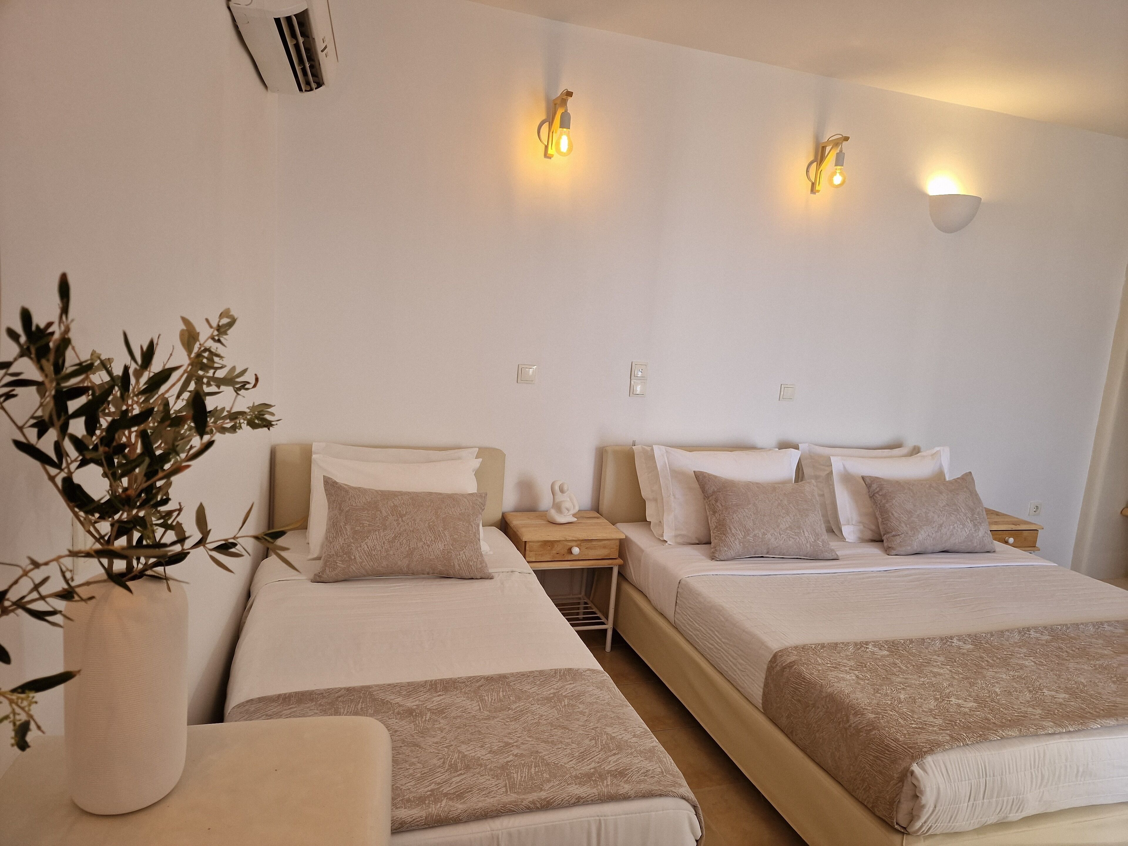 A hotel room with two beds, a bedside table, a nightstand, a potted plant, and wall-mounted lights.