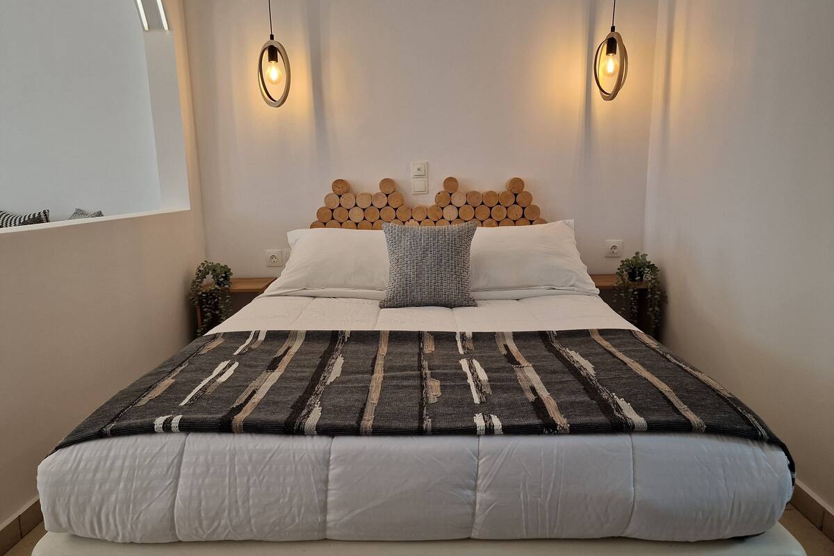 Una cama bien hecha con un edredón a rayas, dos mesitas de noche con plantas, un cabecero de troncos y dos lámparas colgantes.
