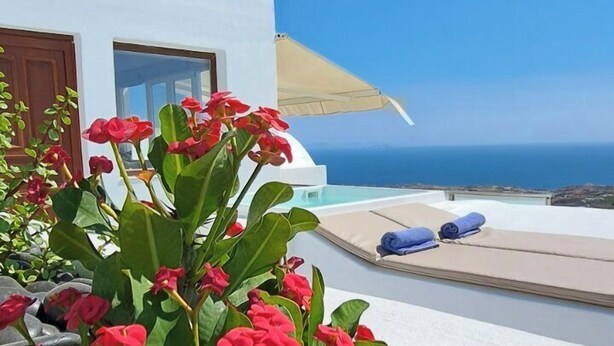 A pool area with a white building, red flowers, and a clear blue sky. - Amerisa Suites & Villa (Santorini)