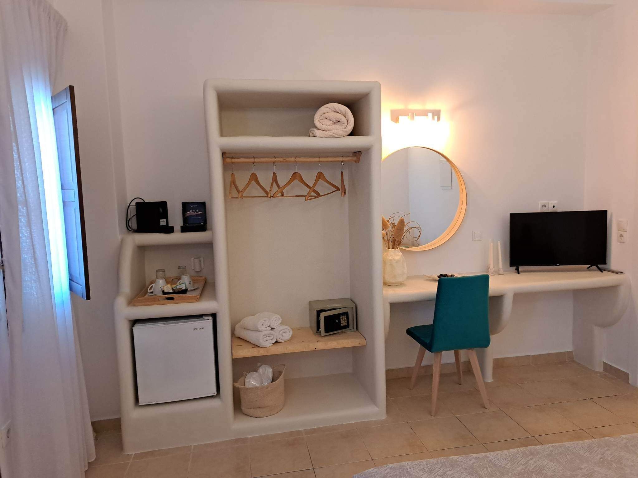 Una habitación de hotel compacta con kitchenette, un escritorio y una silla, televisor, espejo y estante con toallas y un refrigerador pequeño.