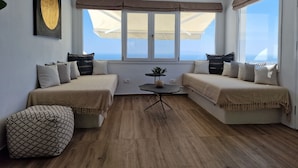 A modern room with two sofas facing a large window, a small table with a plant, and a ceiling light. - Amerisa Suites & Villa (Santorini)