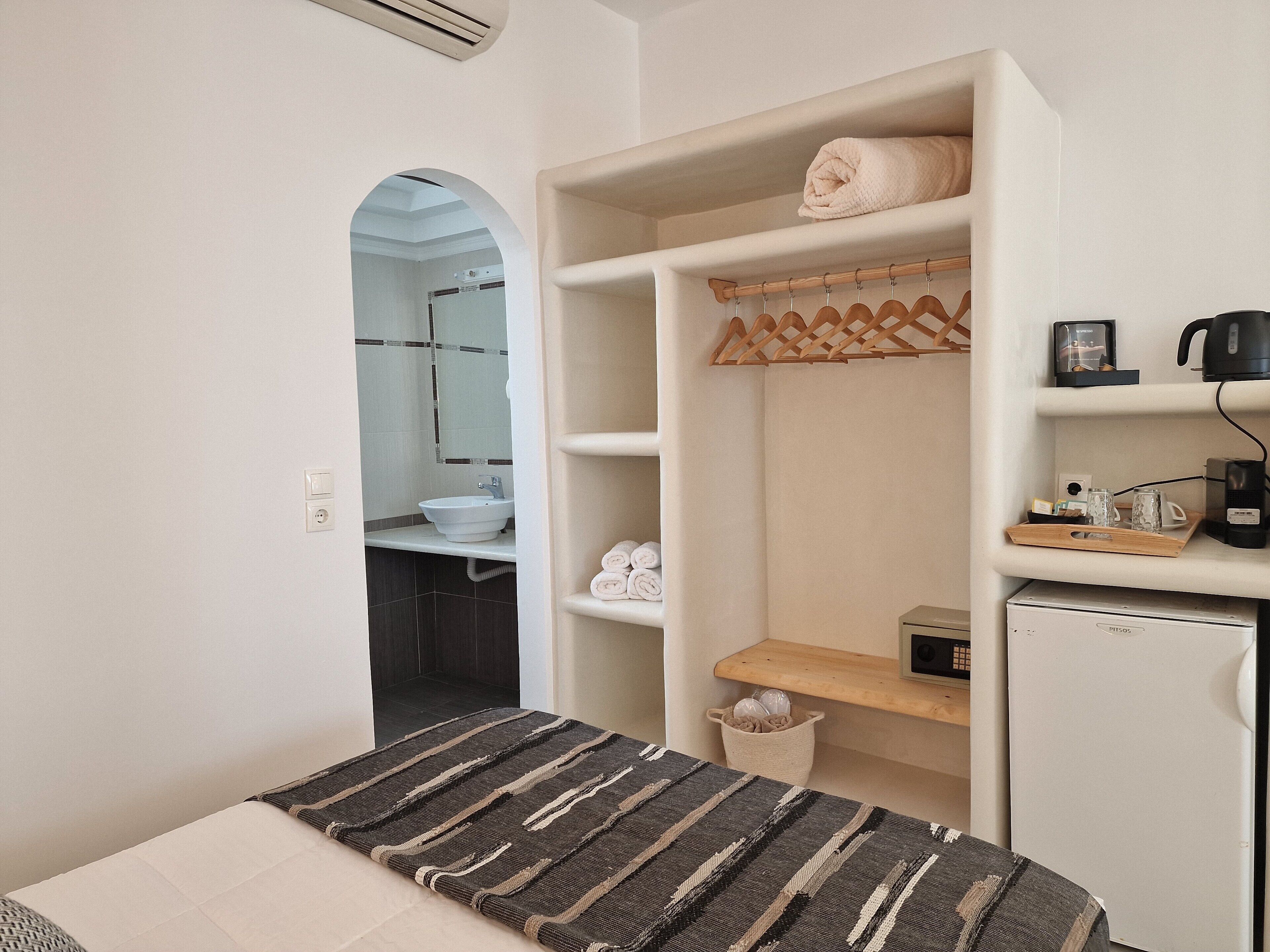 Habitación de hotel compacta con cama, una kitchenette pequeña, un baño visible a través de un arco y un almacenamiento con toallas y artículos de aseo.