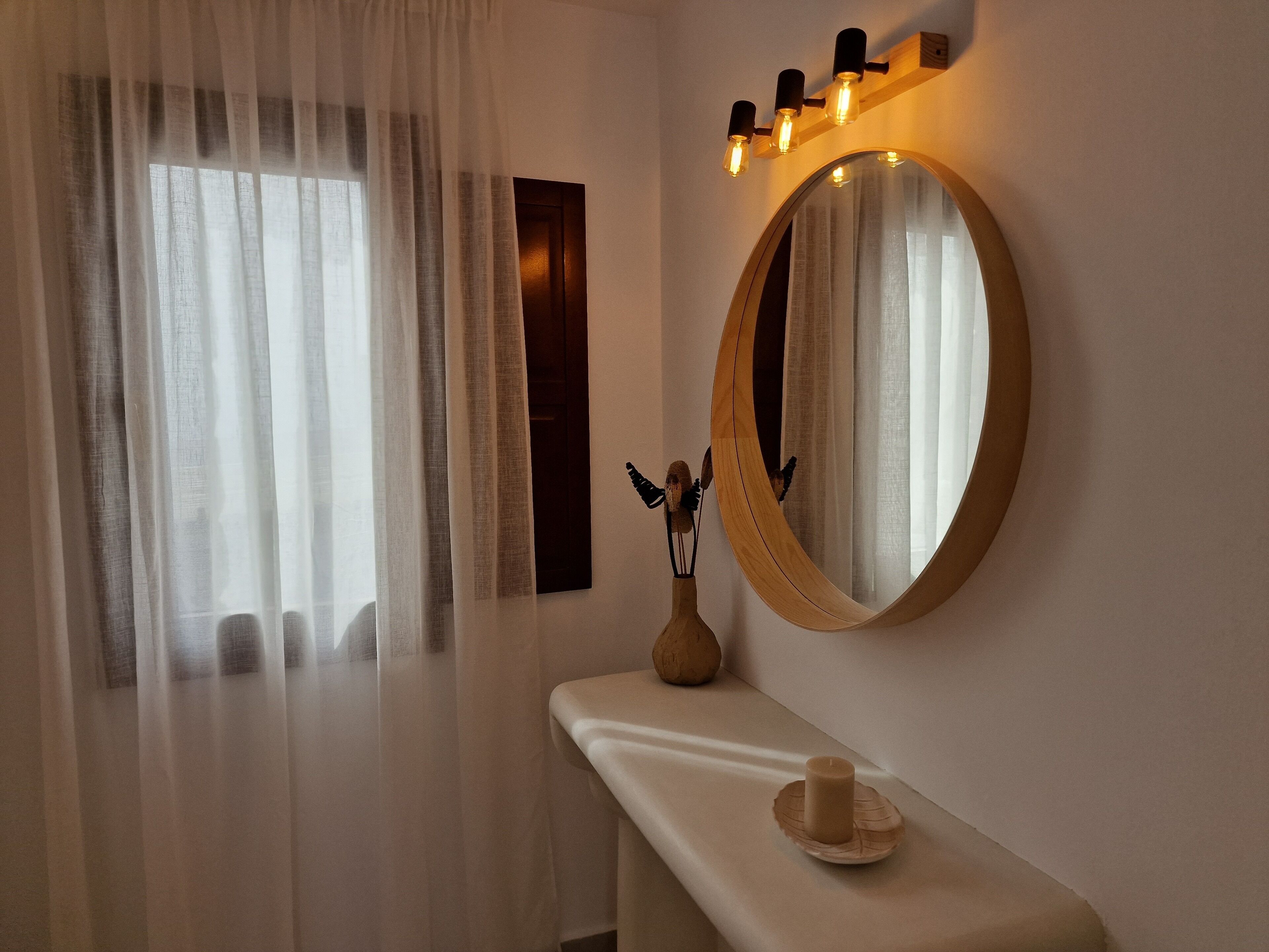Un baño con un espejo grande, un tocador con una vela y una ventana con cortinas traslúcidas.