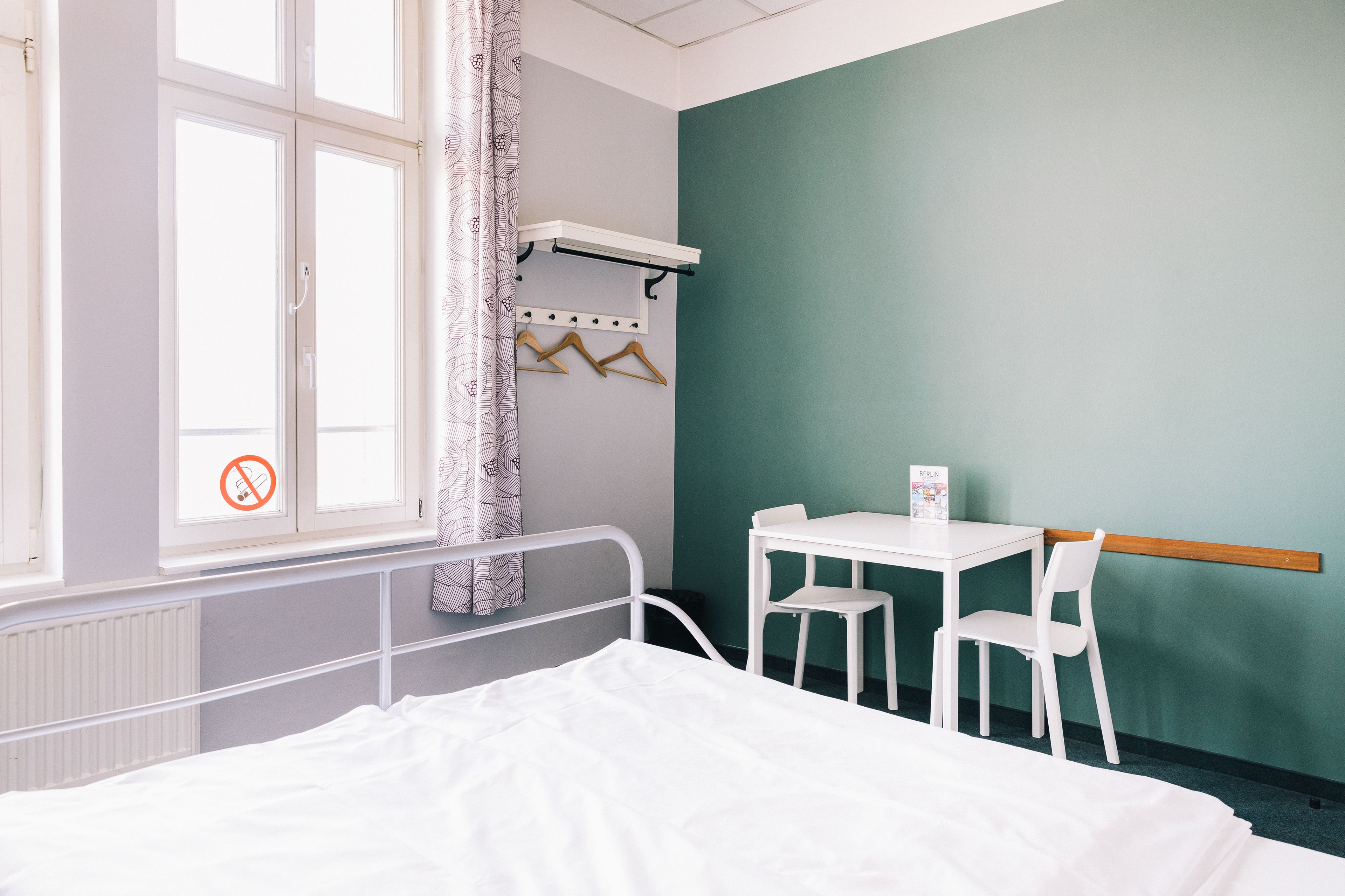 Una habitació d'hospital amb un llit, una taula petita amb dues cadires, una finestra amb cortina i una prestatgeria fixada a la paret amb penjadors.