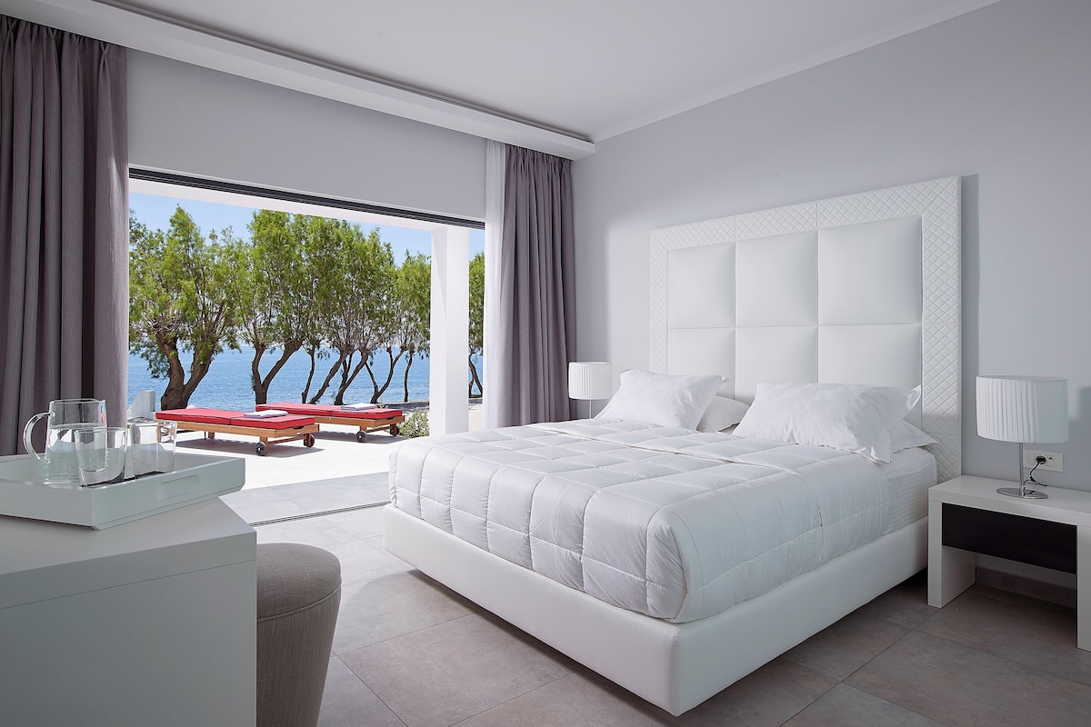 A modern bedroom with a large bed, a bedside table, a lamp, and a view of the sea through large windows.