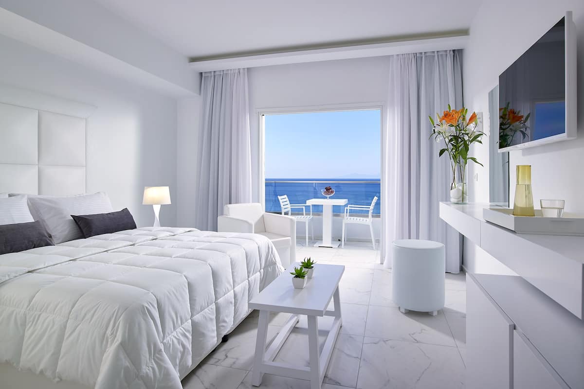 A modern hotel room with a large bed, a balcony with a view of the sea, and a small table with chairs.