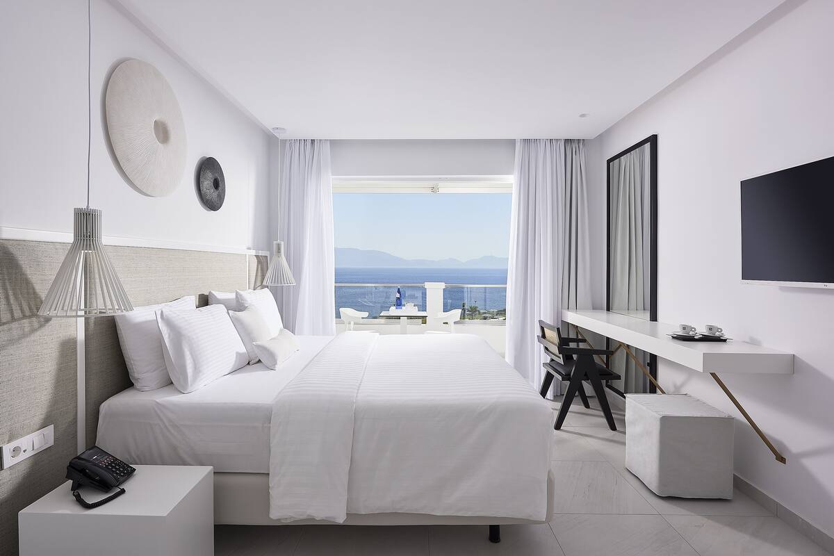 A modern hotel room with a large bed, a desk, a TV, and a view of the sea.