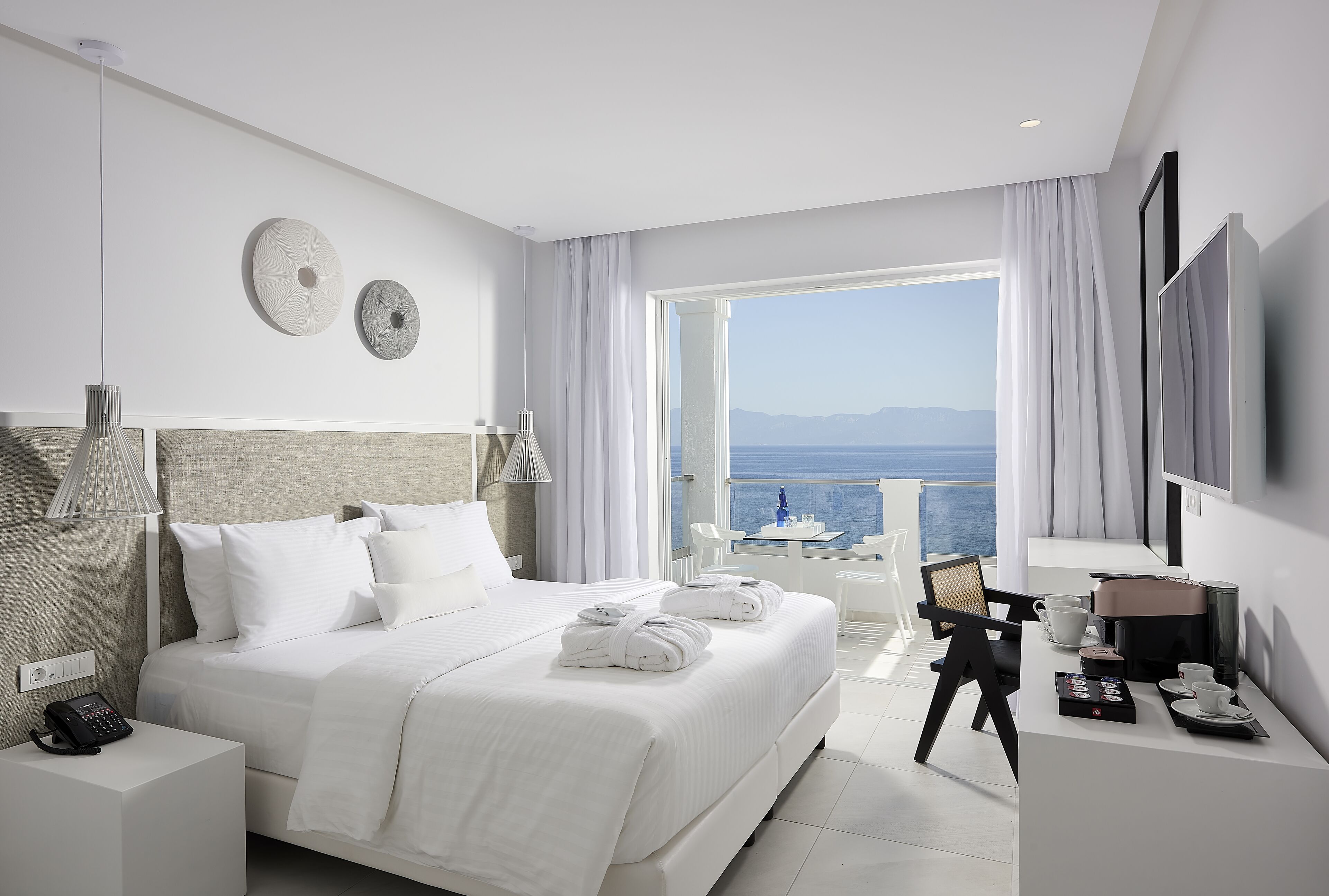 Una habitación de hotel moderna con una cama grande, un escritorio con una computadora portátil, un televisor de pantalla plana y un balcón con vista al mar.