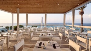 Een restaurant met witte tafels en stoelen, grote ramen met uitzicht op de oceaan en een houten plafond.