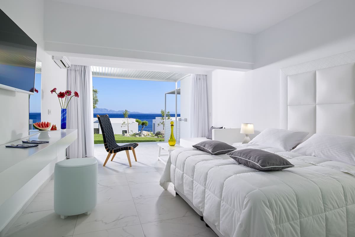 A modern hotel room with a large bed, a chair, a small table, and a view of the sea.