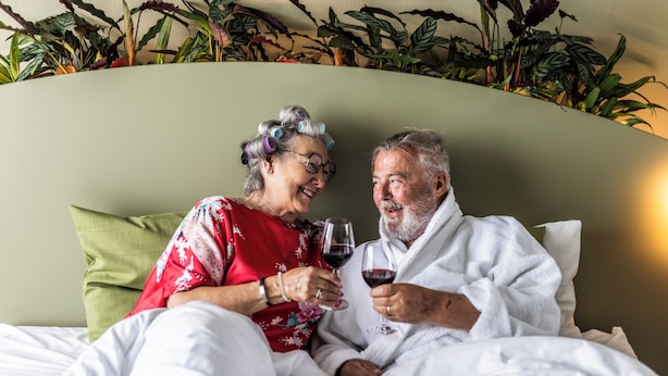 A couple in bed with a green headboard and a plant wall behind them.