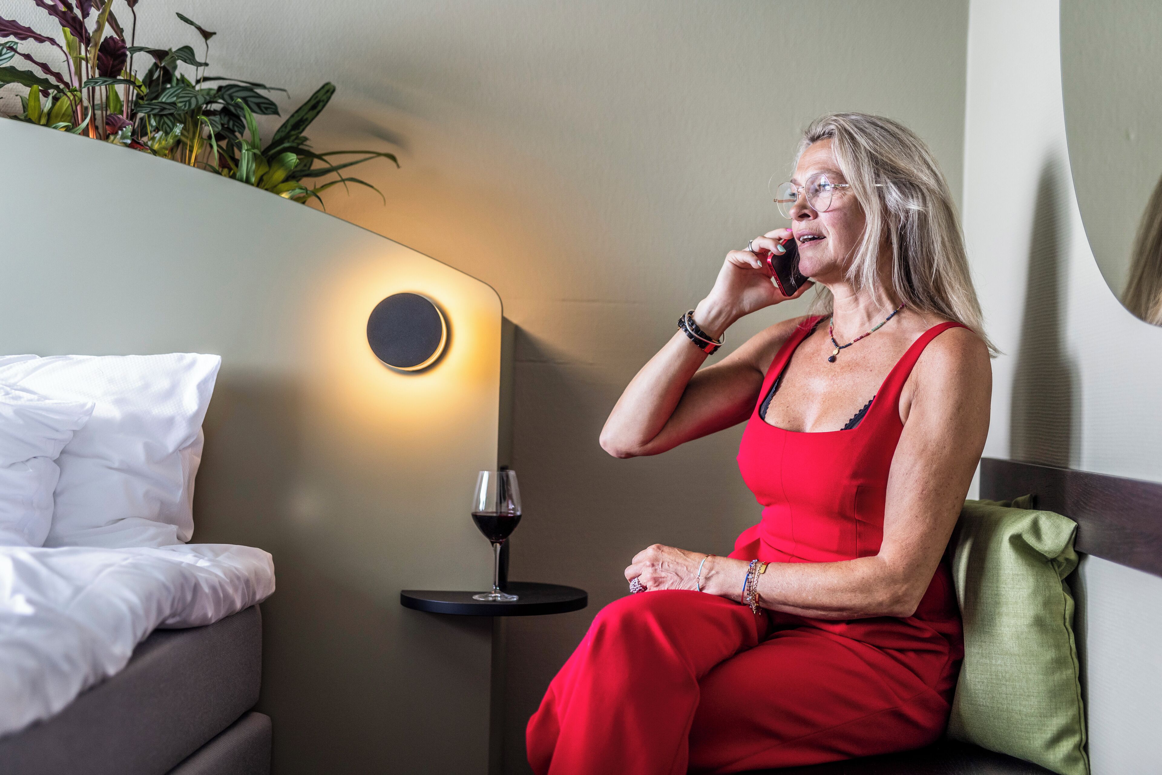 Une femme vêtue d’une robe rouge est assise sur un lit dans une chambre d’hôtel. On aperçoit une table de chevet, un verre à vin et une lampe.