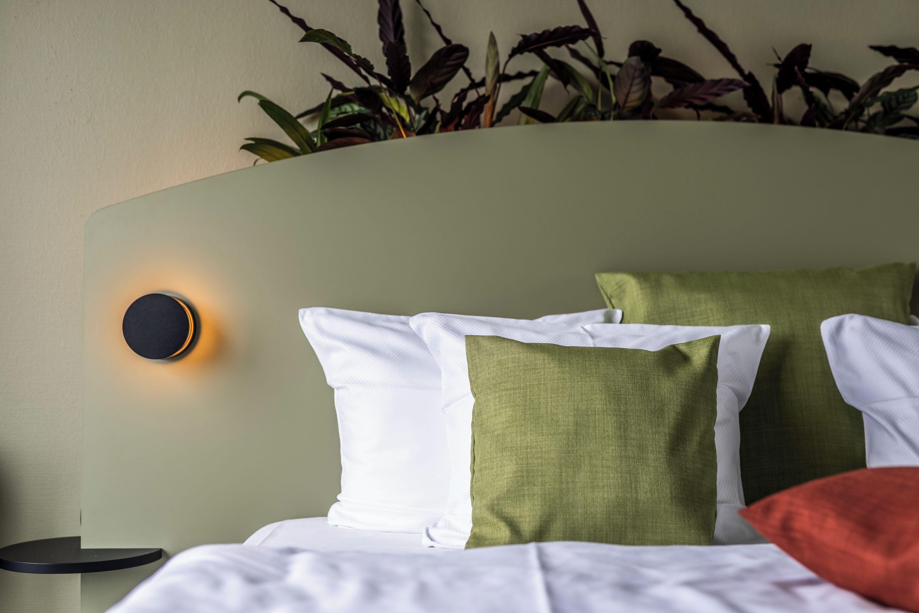 A neatly made bed with white and green pillows, a round wall-mounted light, and a small bedside table.
