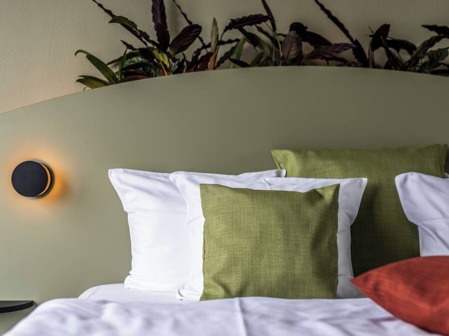 Una cama bien hecha con almohadas blancas y verdes, una lámpara redonda montada en la pared y una mesita de noche pequeña.