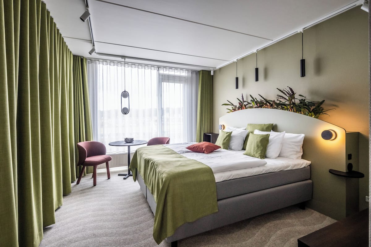 A modern hotel room with a bed, green curtains, a small table, and a chair.