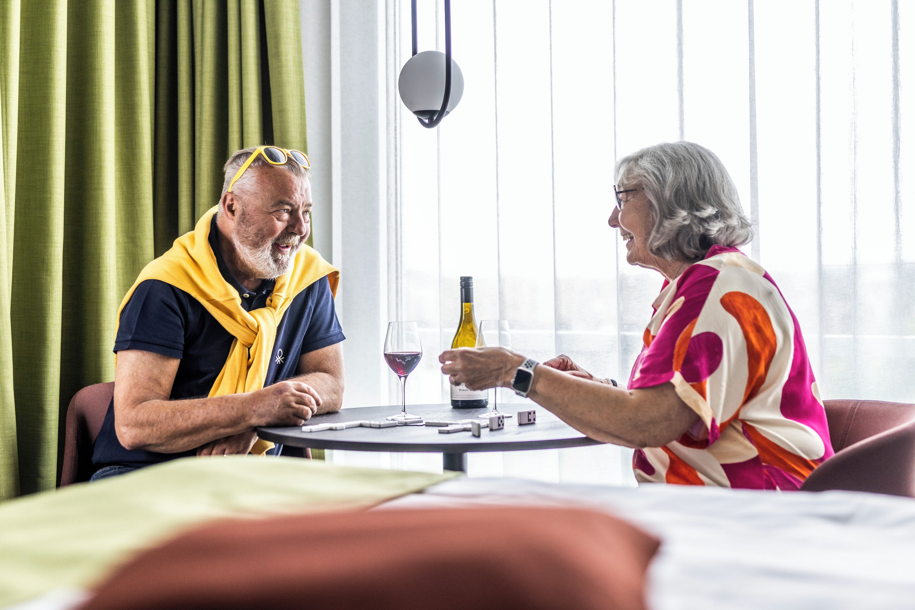 Two people seated at a round table with a bottle of wine and glasses, in a room with a large window and curtains.