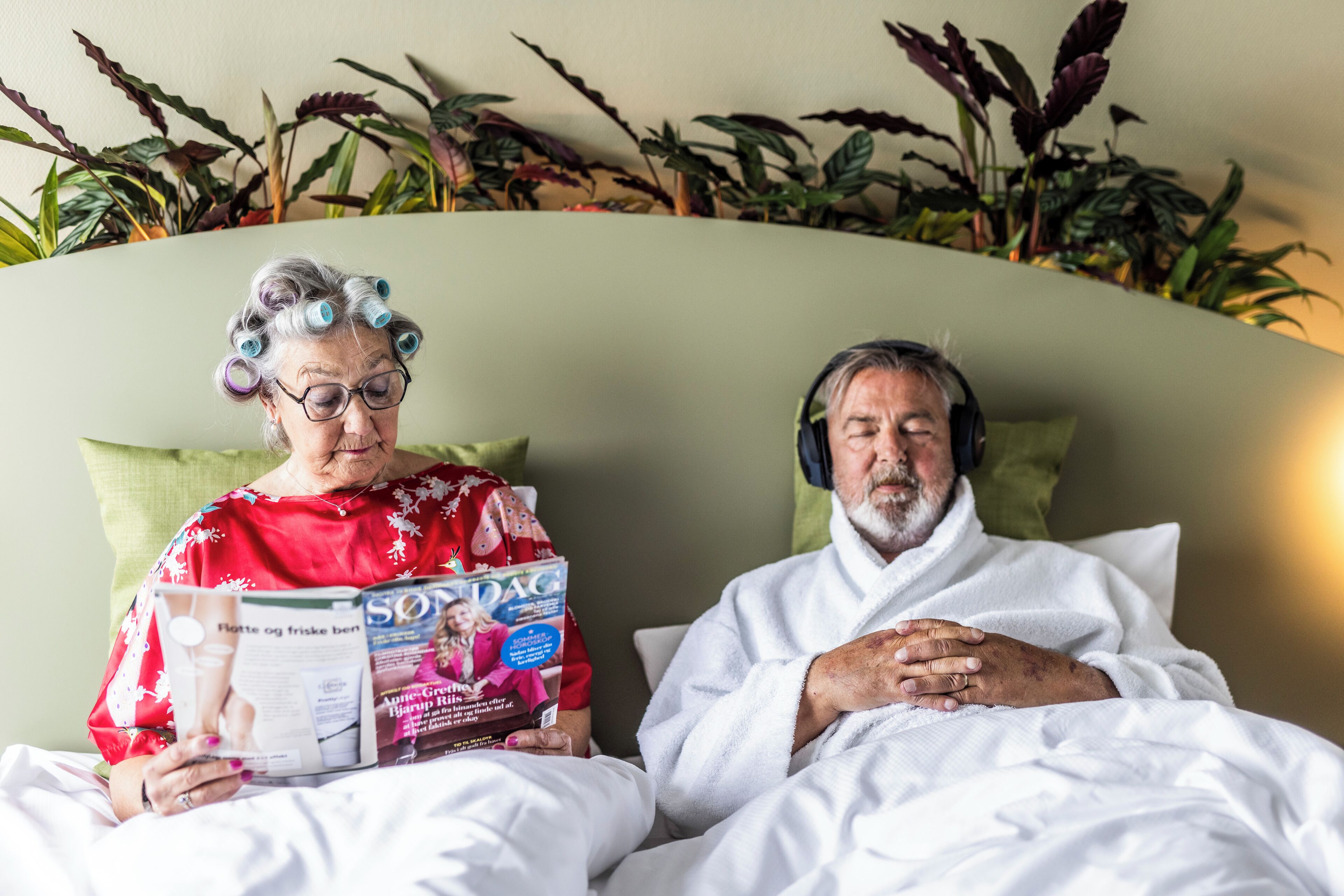 Two people sitting on a bed with a magazine, surrounded by plants.