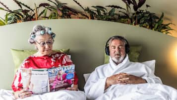Two people sitting on a bed with a magazine, surrounded by plants.